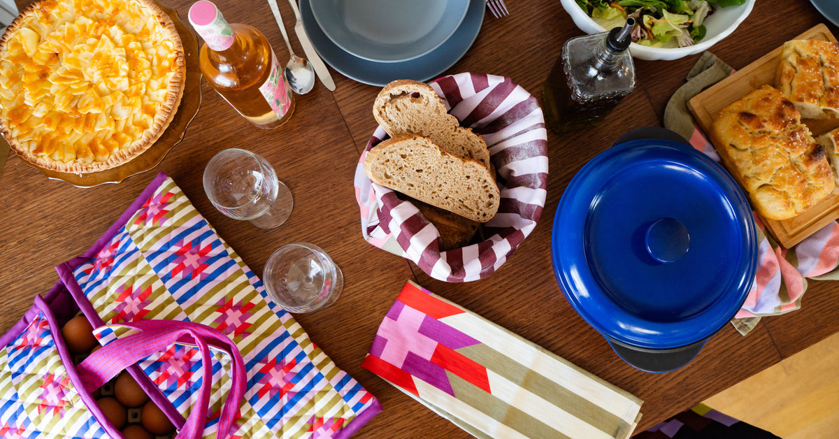 A styled dining table scene with a colorful striped picnic blanket, a blue lidded serving dish, bread, salad, pie, glasses, and a bottle of rosé wine on a wooden table.